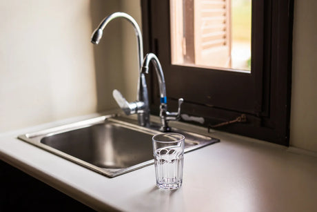 Clean kitchen sink fitted with dual faucets for standard RO and remineralization alkaline RO water, with a glass of fresh filtered water on countertop.