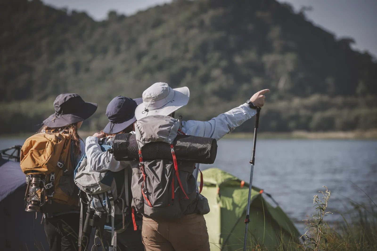 Three hikers with backpacks at a lakeside campsite, preparing to use a water purifier to treat natural water sources