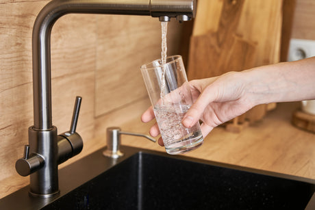Hand filling a clear drinking glass with water from a modern kitchen faucet above a black sink