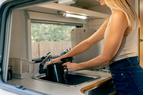 A woman fills a black kettle at a compact RV kitchen sink, demonstrating daily water use in a mobile home.