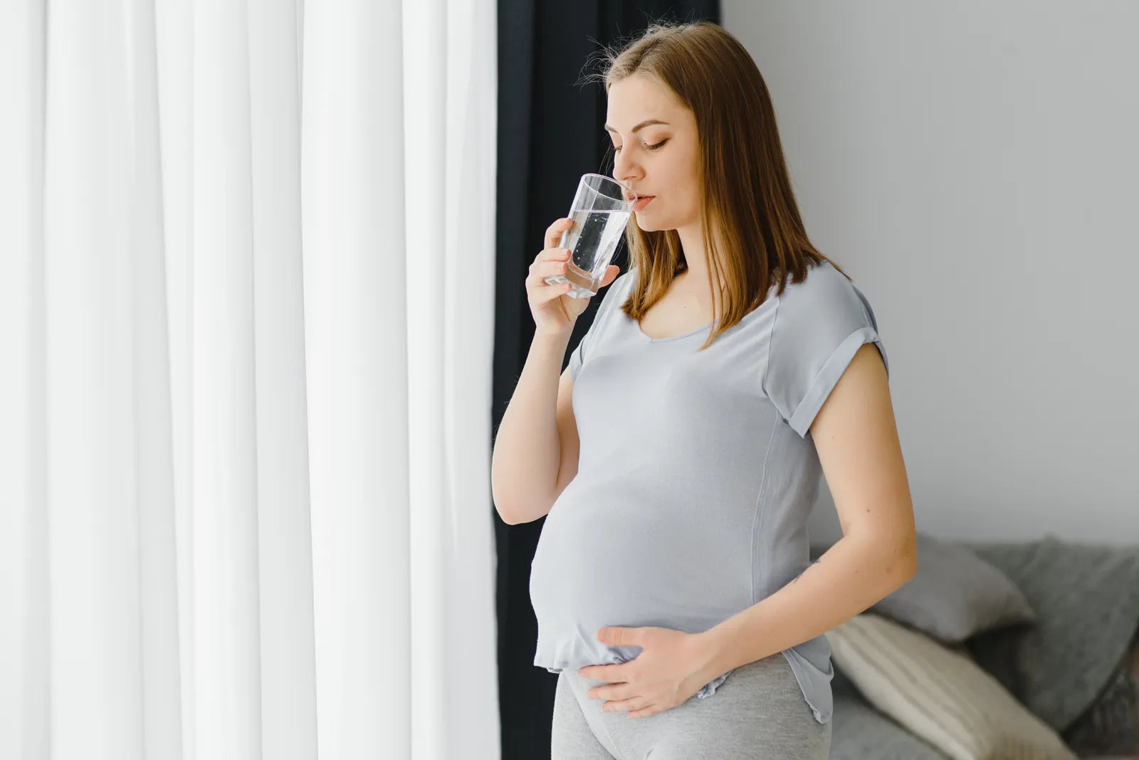 Pregnant woman drinking a glass of clean water while gently touching her belly, demonstrating safe hydration practices during pregnancy for maternal and fetal health.