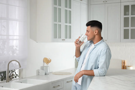 A man drinks a glass of clean water in a bright white kitchen, highlighting safe drinking water benefits in small apartments.