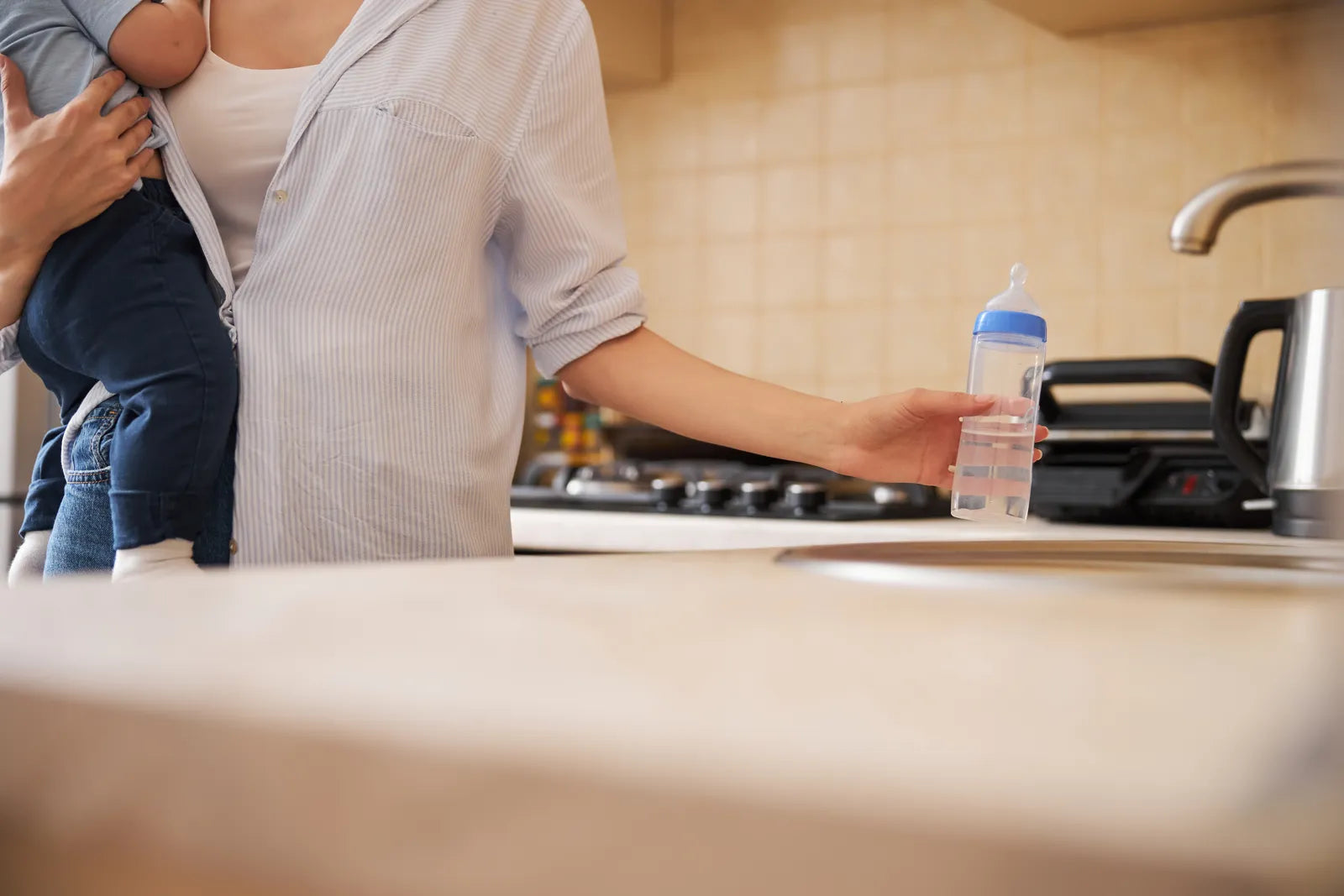 A parent holding a baby and a water bottle in the kitchen, highlighting the scenario of preparing safe drinking water and formula for infants at home.