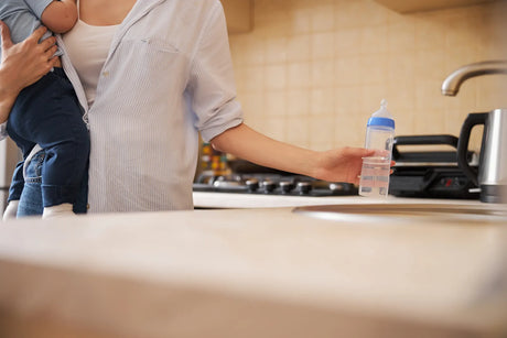 A parent holding a baby and a water bottle in the kitchen, highlighting the scenario of preparing safe drinking water and formula for infants at home.