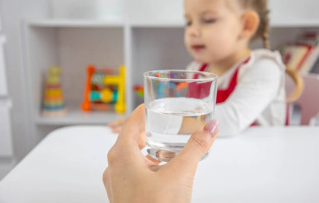 A hand offers a glass of water to a toddler, raising questions about alkaline water safety for babies.