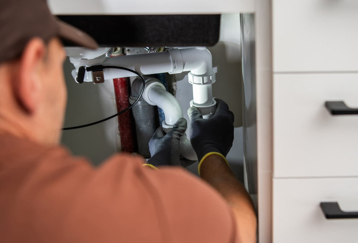 Open kitchen sink cabinet showing water valves, faucet hoses, and drain plumbing.