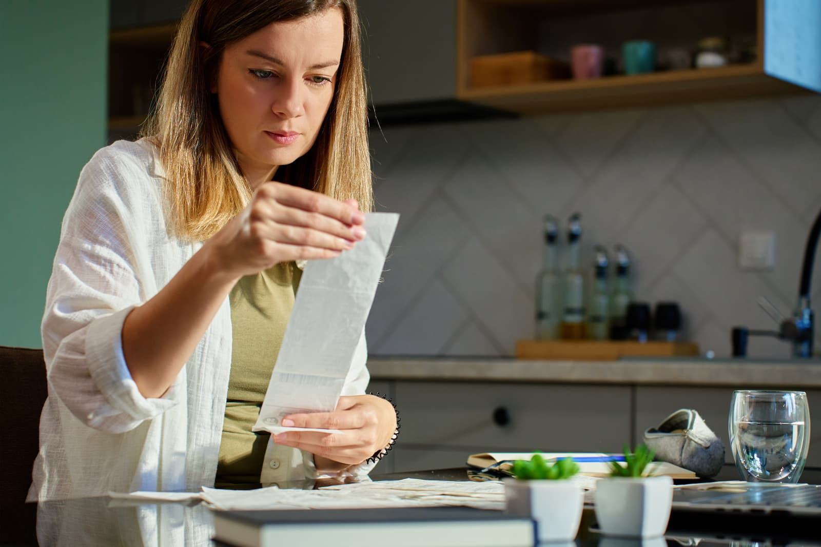 Woman reviewing household expenses at a kitchen table beside a glass of drinking water