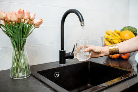 A glass is filled with fresh water from a kitchen faucet beside a sink and fruit bowl.
