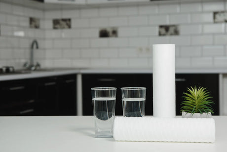 Two new water filter cartridges and glasses of water sit on a kitchen countertop, ready for replacement.