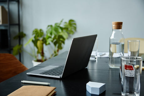 Laptop and water glasses on a workspace table beside a clear water carafe and indoor plants.