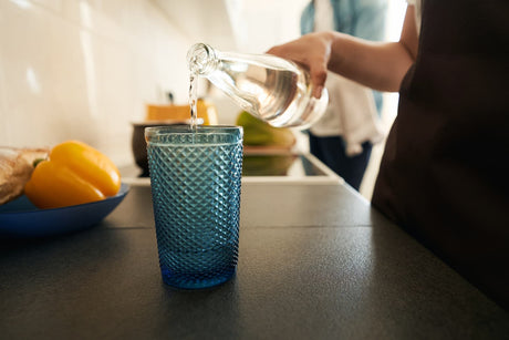 Person pouring clear drinking water from a glass bottle into a blue tumbler