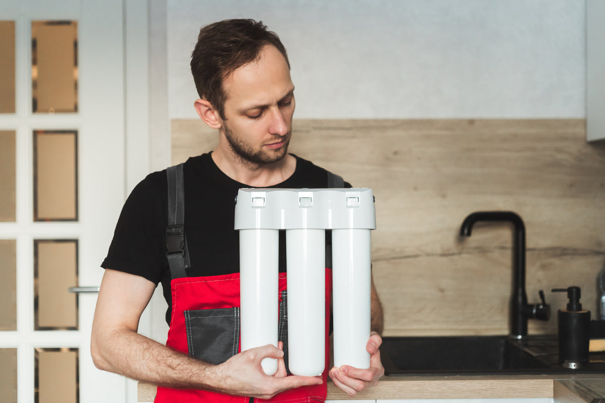 Man holding a three-cartridge water filtration system in a kitchen before under-sink installation