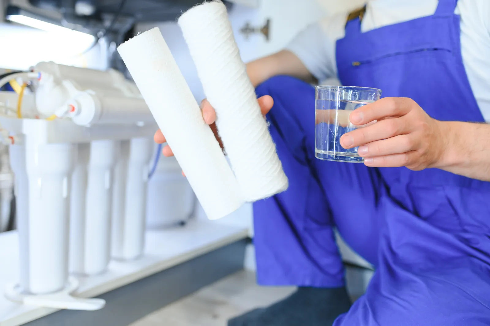 A technician in blue overalls holds new white water filter cartridges and a glass of clean water, demonstrating the result of a sanitized water filter system.