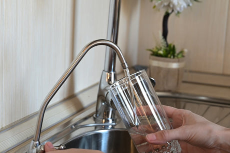 A person filling a clear drinking glass with water from a separate filtered water faucet above a kitchen sink