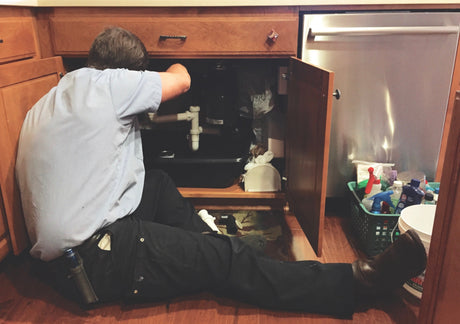 A person inspects plumbing connections and drain pipes inside an open kitchen sink cabinet.