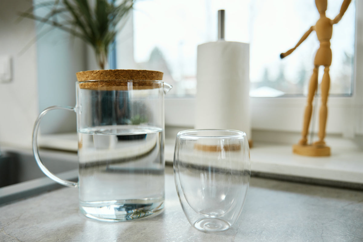 Clear glass pitcher filled with water beside an empty drinking glass on a bright kitchen countertop