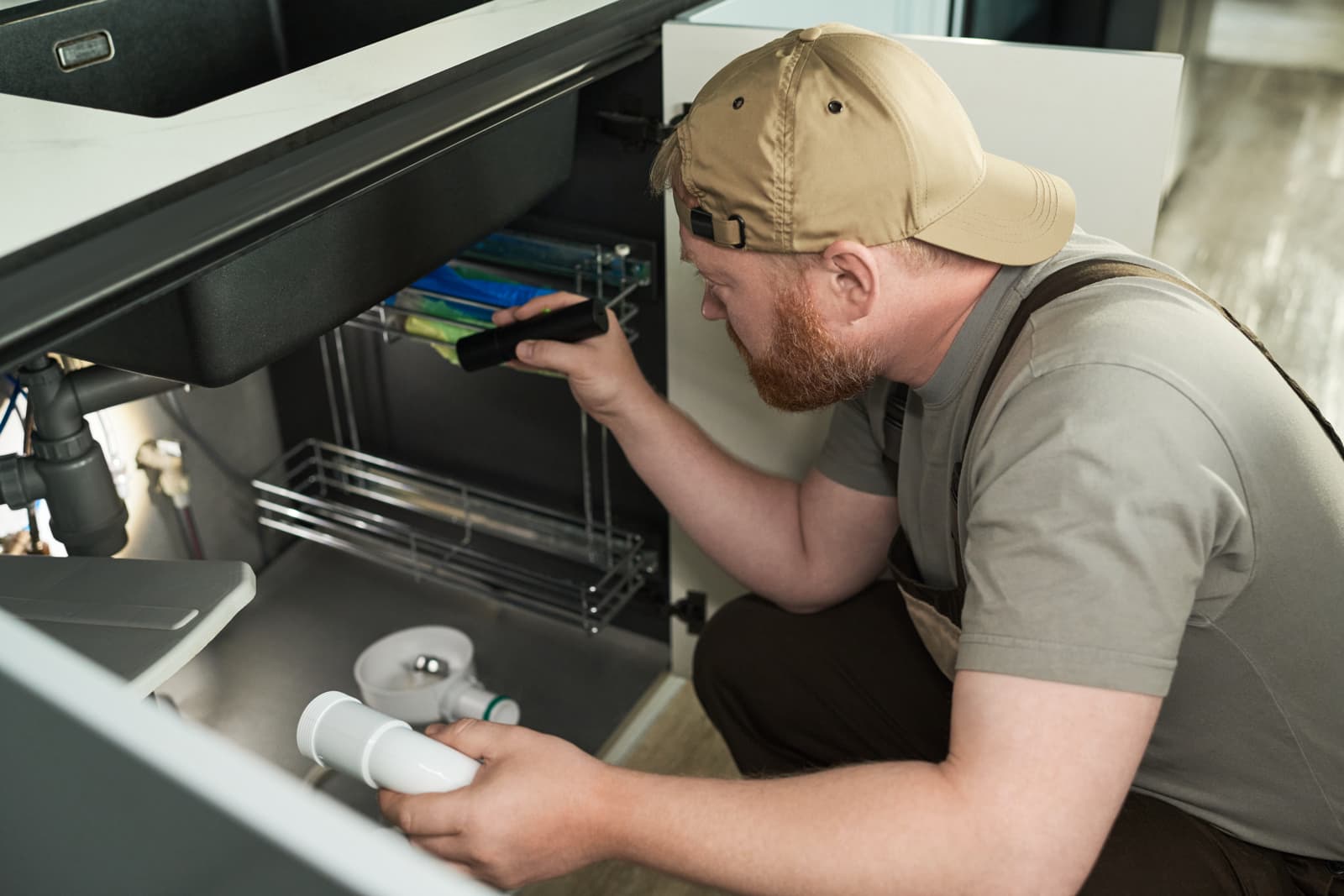 Plumber using a flashlight to inspect under-sink plumbing while holding a drain pipe fitting.