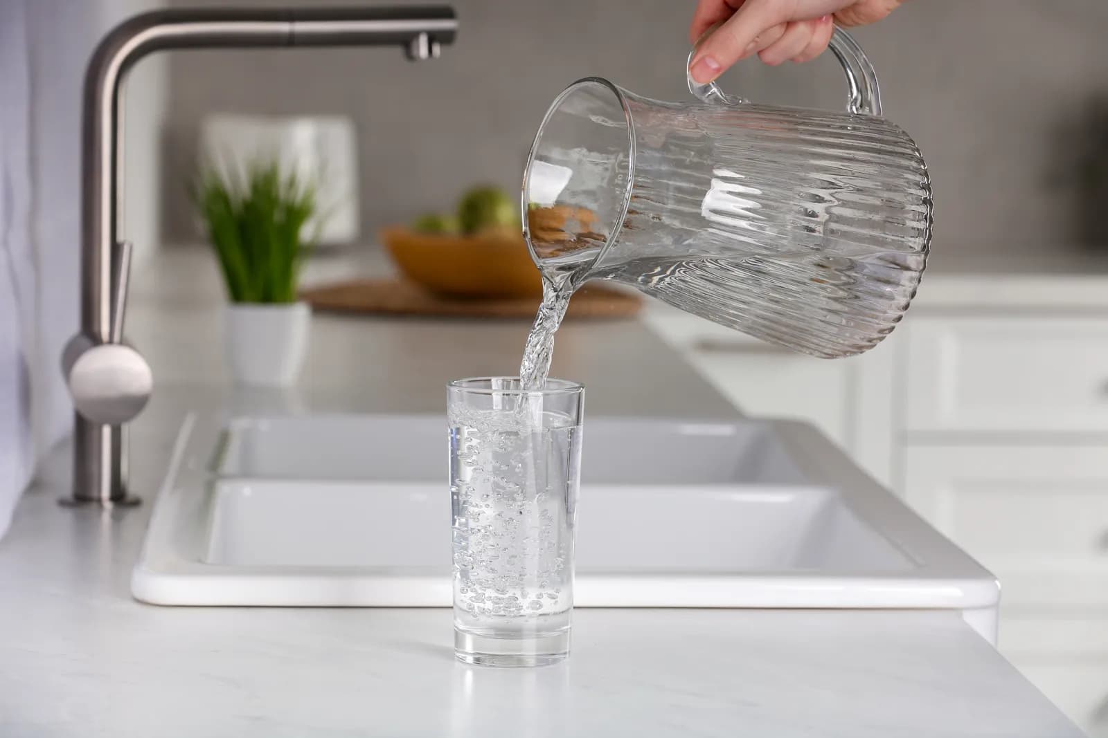 Clear drinking water is poured from a glass pitcher into a cup beside a modern kitchen sink.