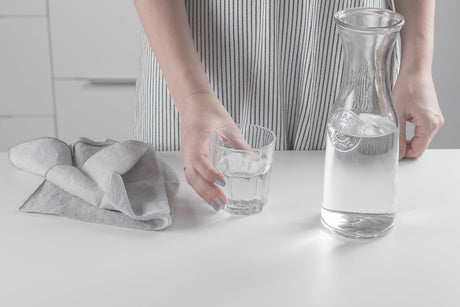 A person holding a glass of drinking water beside a clear glass carafe on a kitchen counter