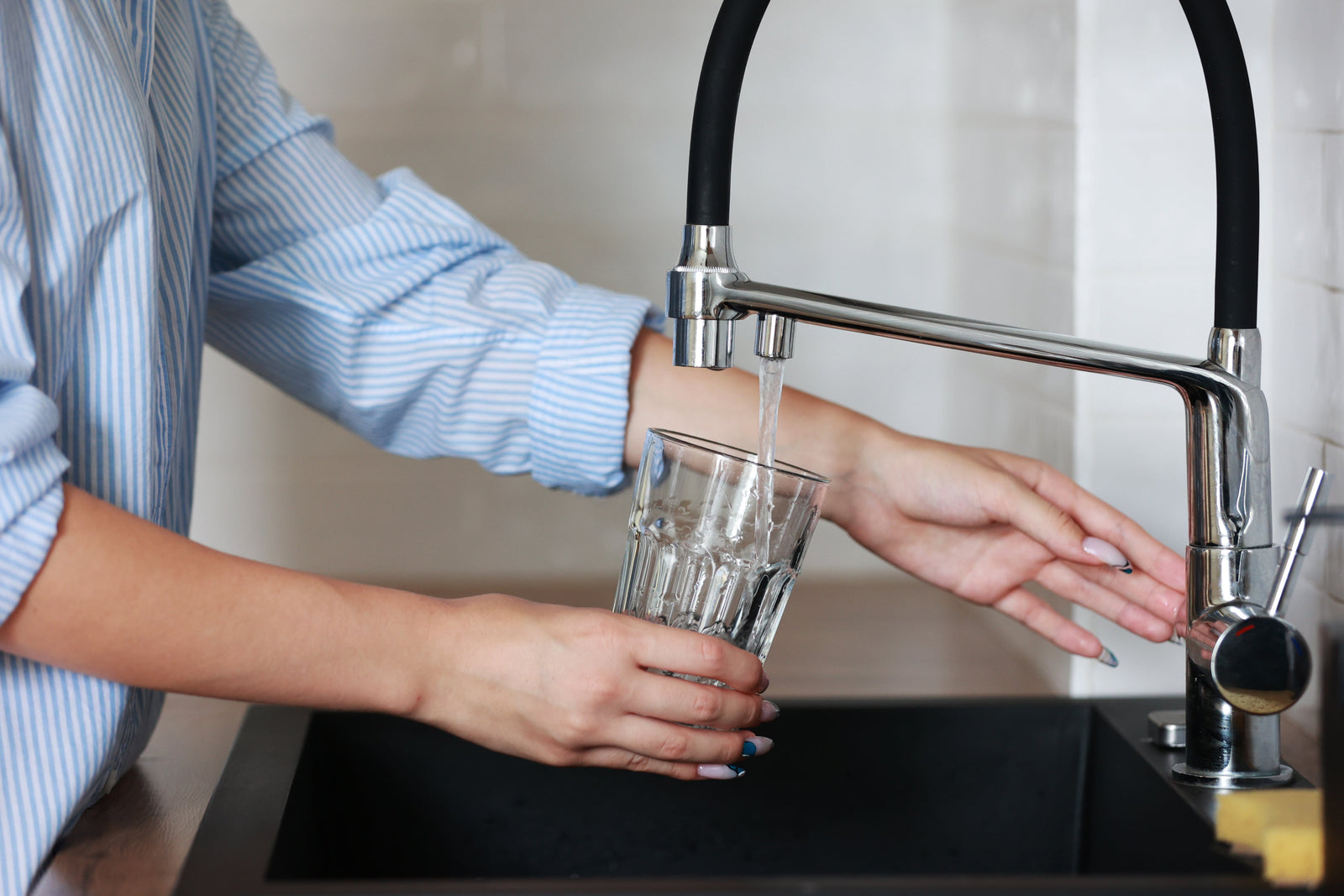 Person filling a clear drinking glass from a kitchen faucet over a black sink.