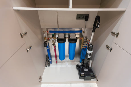 Under-sink water filtration setup with blue housings, valves, plumbing lines, and nearby power outlets.