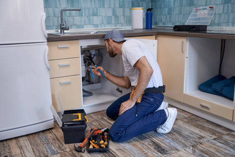 Plumber using pliers to work on pipe connections inside an open kitchen sink cabinet.