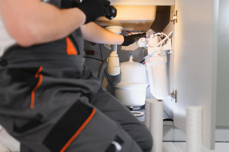 Homeowner inspecting an under-sink water filtration system beside a kitchen sink.
